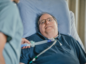 Male patient lying in hospital bed with a Vocolair speaking valve in line 